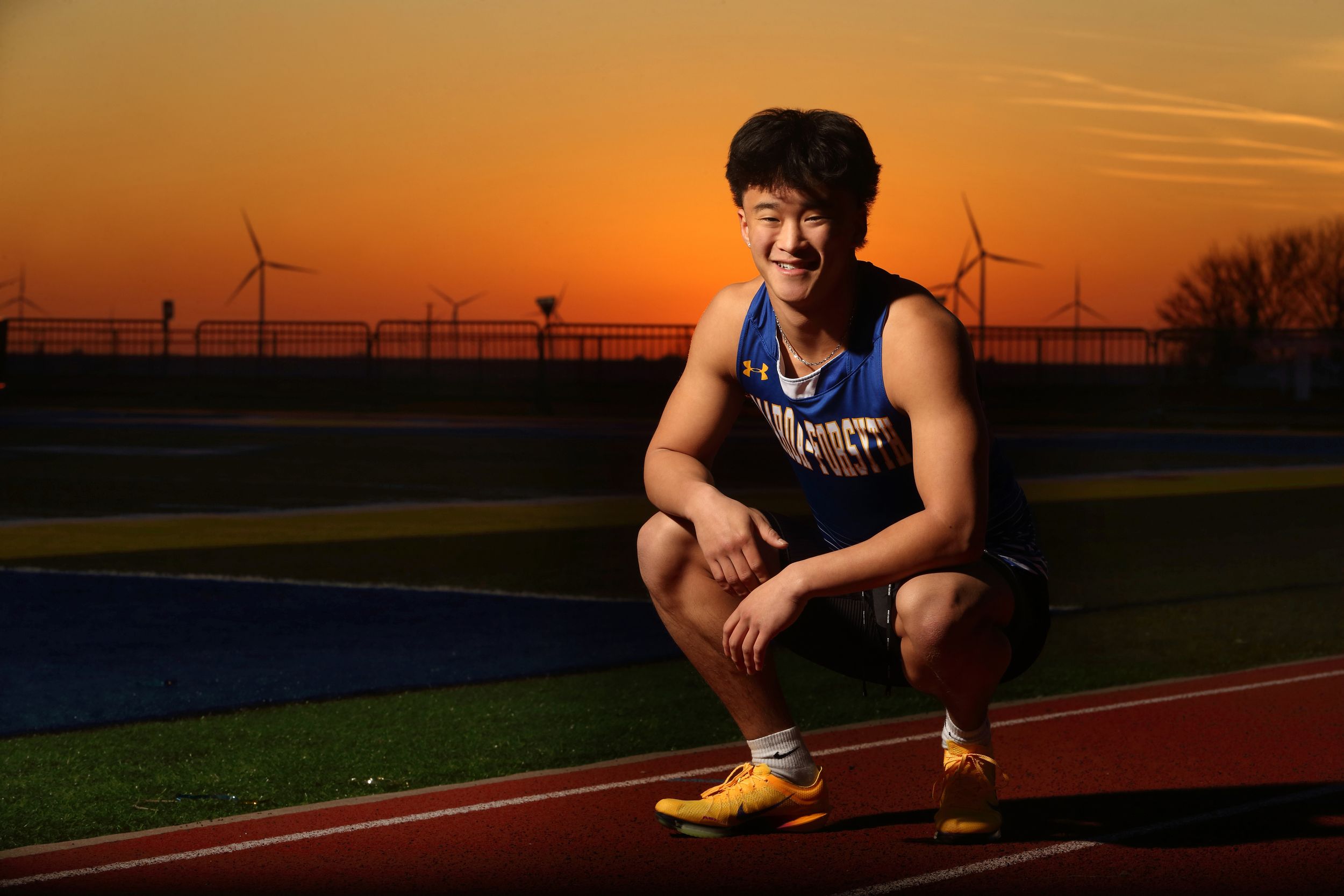 Young Asian male athlete in blue track uniform crouches on running track at sunset with wind turbines in background.