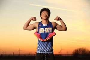 Young athlete in Maria-Forsythe track uniform flexes biceps holding pink shoes at sunset.