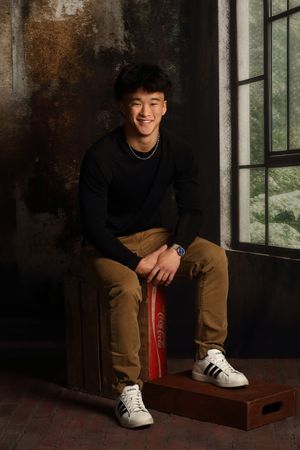 Smiling young Asian man in black sweater and khaki pants sitting on a crate near a window indoors.