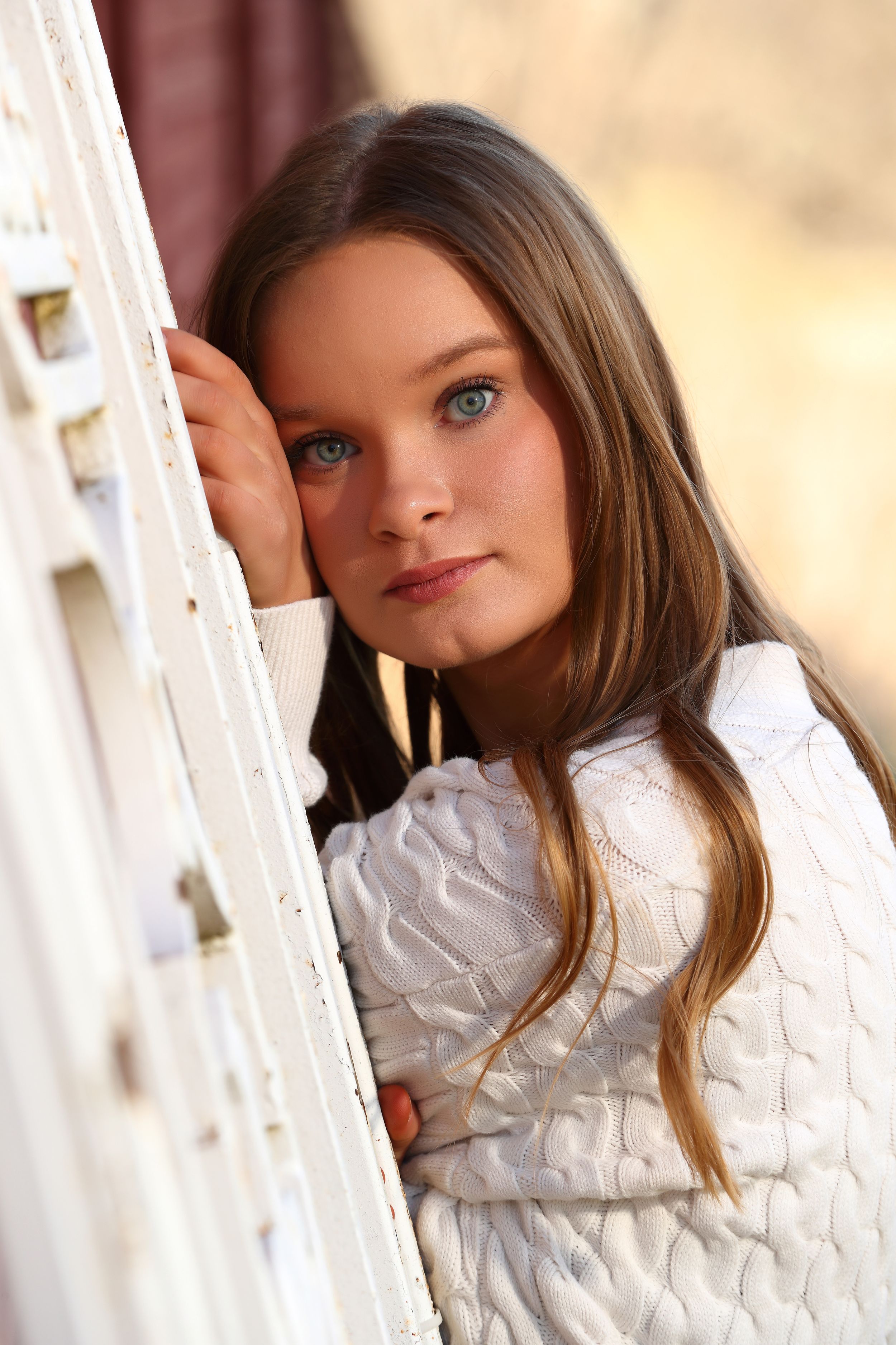 Young girl with blue eyes and long brown hair leans against white shutters wearing a white knit sweater.