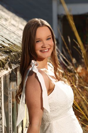 Young woman in white dress smiling outdoors near rustic fence with golden grasses in warm sunlight.