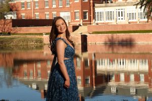 Young woman in blue floral dress posing by a reflective pond with a red brick building backdrop.