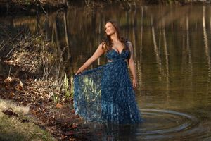 A woman in a flowing blue lace gown stands gracefully in a reflective pond surrounded by autumn foliage.