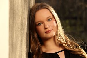 Young blonde woman with blue eyes leaning against a wall, wearing a black top in warm golden light.