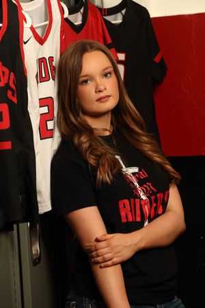 Young woman in a black Riders t-shirt poses confidently in a locker room with red and white sports jerseys hanging behind her.