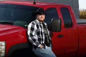Man in black and white flannel shirt and cowboy hat leaning against a red Chevrolet pickup truck outdoors.