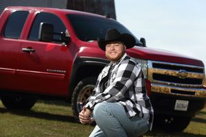 Man in black cowboy hat and plaid shirt crouching beside a red Chevrolet Silverado pickup truck outdoors.