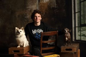 Young man in black graphic tee sits on chair flanked by two small dogs in moody studio portrait.