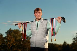 Young male golfer proudly displays colorful medals on a golf club resting across his shoulders outdoors.