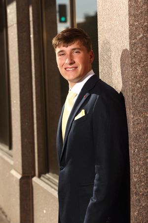 Young man in navy suit with yellow tie and pocket square smiling outdoors.