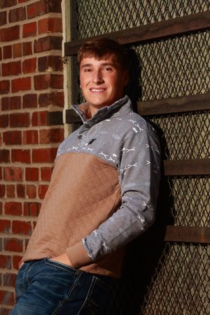 Smiling young man in gray patterned pullover and jeans leaning against brick wall with metal grate backdrop.