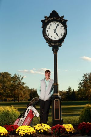 Young man posing by South Side CC clock with golf bag amid colorful flowers.