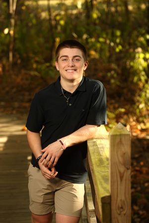 Smiling young man in black polo and khaki shorts leaning on wooden railing outdoors in autumn setting.