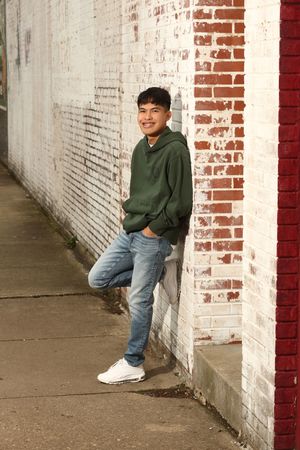 Someone in a green hoodie and jeans leans casually against a white and red brick wall in an urban setting.
