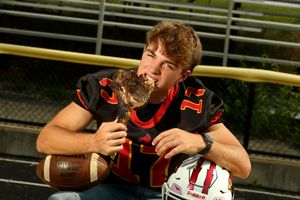 Teen football player in black jersey kisses trophy while holding football and helmet in stadium.