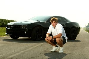Young man crouching in front of a sleek black Chevrolet Camaro on an open road.