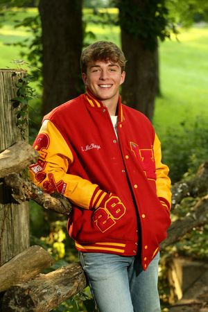 Smiling teen in red and yellow varsity letterman jacket leaning on rustic wooden fence outdoors.