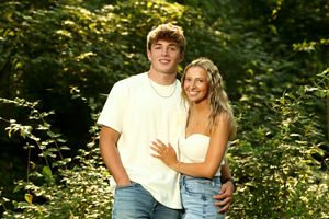 Young couple smiles together outdoors surrounded by lush green foliage in natural setting.