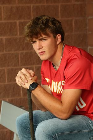Young male athlete in red jersey holds baseball bat, wearing smartwatch, seated against brick wall.