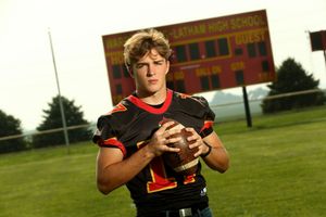 Young football player in black jersey holding a football on a field near a high school scoreboard.