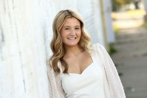 Smiling young woman in white dress leaning against a white brick wall outdoors.
