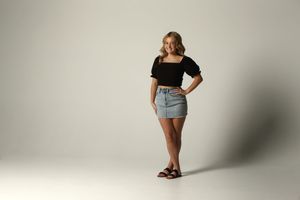 Young woman in black top and denim mini skirt posing confidently in a studio setting.