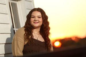 Young woman with long curly red hair smiling outdoors at golden sunset, wearing a brown top and tan cardigan.