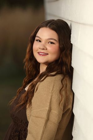 Young woman with long wavy brown hair smiles warmly, leaning against a white wall in a tan knit cardigan.