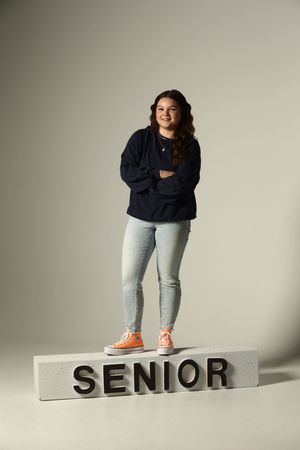 A smiling high school senior stands confidently on a block reading SENIOR, wearing a navy sweatshirt, jeans, and orange Converse.