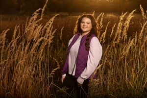 Young woman in purple vest smiling in golden sunlit tall grass field during sunset portrait photography.