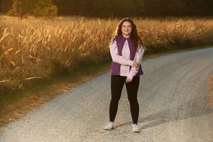 Young woman in purple vest and black leggings standing on a rural gravel road beside golden wheat fields at sunset.