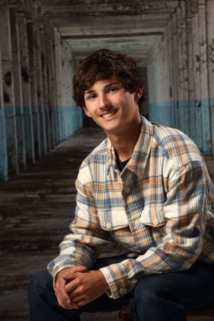 Portrait of someone in a plaid button-down shirt sitting in an industrial corridor with blue walls.