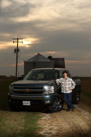 A pickup truck sits parked against a dramatic sunset sky with utility poles and grain bins silhouetted in the background.