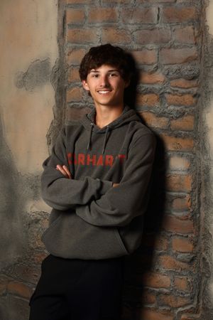 A person wearing a gray Carhartt hoodie smiles while leaning against a brick wall in a portrait-style photo.