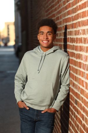 Young man in sage green hoodie smiling against a red brick wall outdoors.