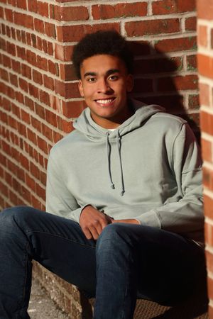 Smiling young man in a light gray hoodie and jeans sitting against a red brick wall in sunlight.