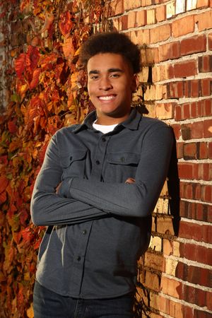 Smiling young man with arms crossed leaning against a brick wall with colorful autumn foliage in fall.