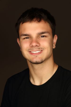 Young man with short brown hair smiling confidently, wearing a black t-shirt against a dark background.