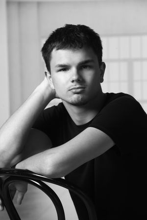 Black and white portrait of a young man in a black t-shirt leaning on a chair with hand raised to his head.