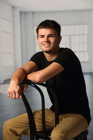 Young man in black t-shirt and khaki pants smiling while sitting on a black chair in a bright studio.