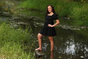 A person in a black dress stands in shallow water near grassy banks on a summer day.