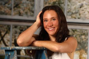 A person with dark hair leans casually on furniture and smiles warmly while wearing a light colored top.