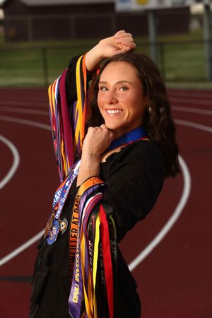 Graduate with colorful academic ribbons and medals poses by running track wearing black graduation attire.