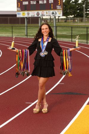 Student athlete displays collection of medals and trophies while standing on red running track wearing formal attire.