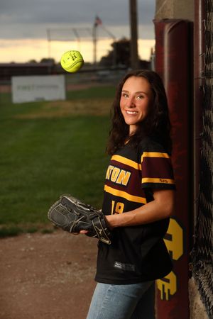 Softball player in black team jersey holds glove and ball while leaning against dugout post at sunset.