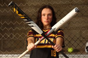 Softball player in black and yellow uniform poses with crossed bats against chain link fence backdrop.