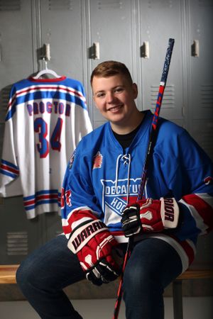 Hockey player poses with equipment in front of team jersey in locker room.