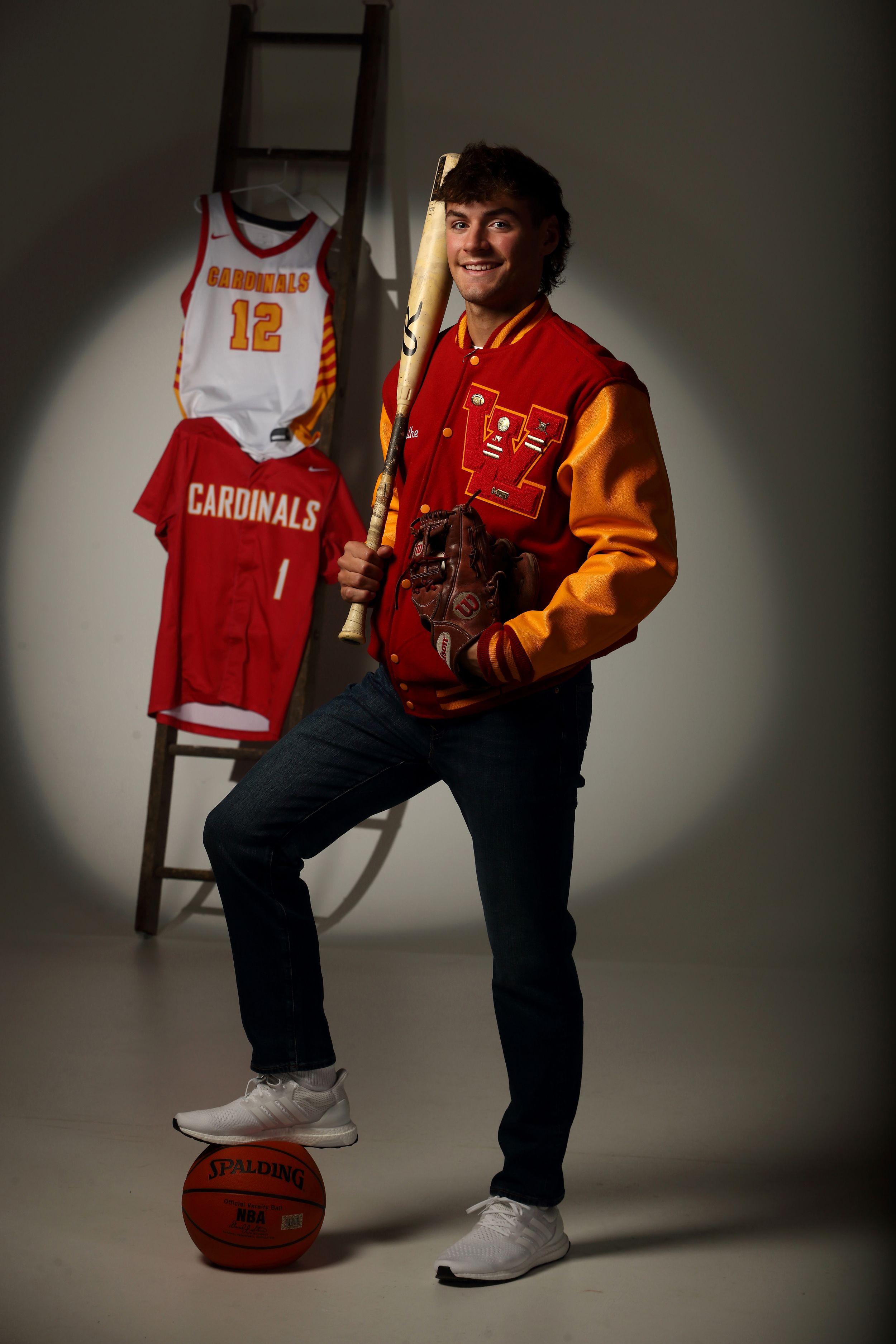 Young man in Cardinals letterman jacket poses with baseball bat, glove, and basketball near sports jerseys on ladder.