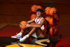 Young male basketball player poses confidently in red jersey surrounded by Wilson basketballs in a gym.