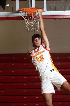 A Cardinals basketball player wearing jersey number 12 hangs from a basketball hoop in a red gymnasium.